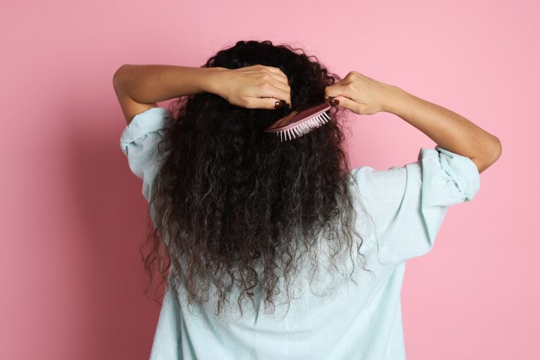 Woman brushing her curly hair on pink background, back view