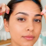 Épilation des sourcils au fil réalisée avec précision, mains tenant un fil torsadé près du sourcil