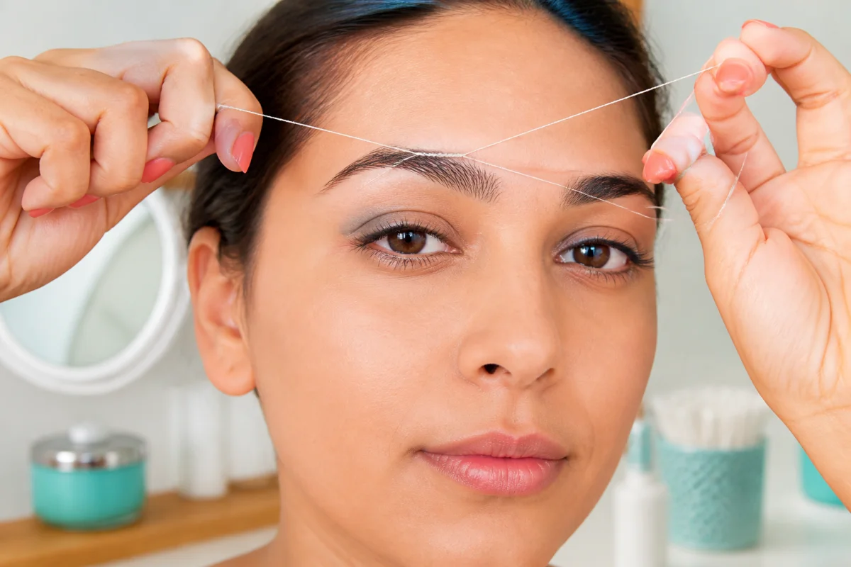Épilation des sourcils au fil réalisée avec précision, mains tenant un fil torsadé près du sourcil
