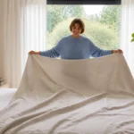 Personne souriante installant une grande housse de couette en lin sur un lit king size dans une chambre lumineuse.